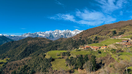Obraz premium A charming rural village with red-tiled roofs nestled in lush green hills, set against the majestic snow-capped Picos de Europa under a clear blue sky. A peaceful, breathtaking view.