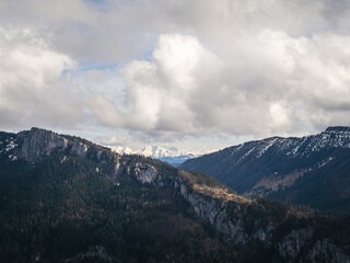 A scenic Bavarian view of a forested mountain ridge with patches of snow, framed by a dramatic cloudy sky and distant snow-capped peaks in the background.