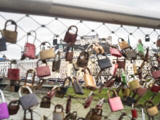 A close-up of colorful love locks attached to a bridge fence in Salzburg, Austria, with a blurred cityscape and river in the background.