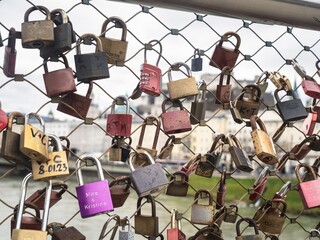 A close-up of colorful love locks attached to a bridge fence in Salzburg, Austria, with a blurred cityscape and river in the background.