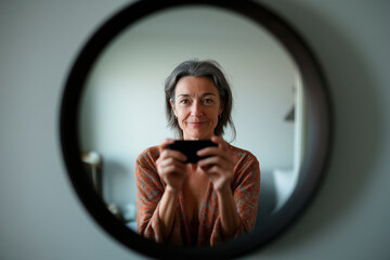 Attractive middle-aged woman in casual dress taking a photo of herself in a mirror in her bedroom in a realistic style for social media