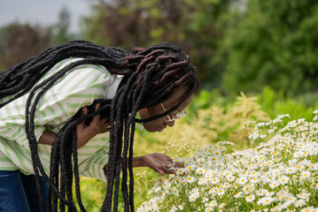 Young botanist with dreadlocks and glasses bending over and smelling chamomile flowers while...