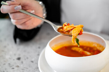 A close-up of a hand holding a spoonful of flavorful soup, featuring vibrant ingredients, served in a modern white bowl. The setting highlights a casual dining experience.