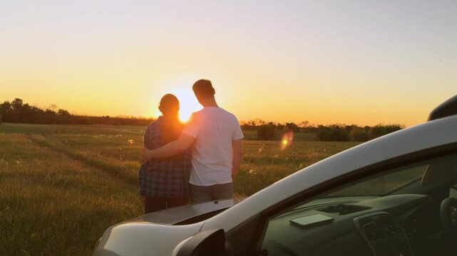 Married family couple in love hugging at sunset by car, romantically spending quiet time together, relaxing outside car. Husband and wife, spouses hugging, standing in field by car. Road trip, travel