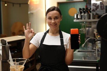 Smiling cafe worker with payment terminal showing OK gesture indoors