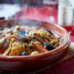 Traditional Moroccan tagine with vegetables, dried fruits, chickpeas served in authentic clay pot. Steaming hot dish photographed in natural light with selective focus. Mediterranean cuisine concept