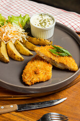 A plate features crispy breaded fish alongside baked potato wedges and a fresh salad. A small bowl of dipping sauce adds flavor