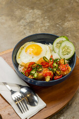 Indonesian sambal matah, A bowl of rice features a fried egg, sliced red chili, and cucumber, arranged attractively on a wooden plate in a warm eatery