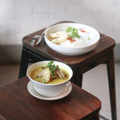 Tasty bowl of soup with fish and greens alongside rice and a side of condiments on rustic wooden stools in a warm, inviting cafe