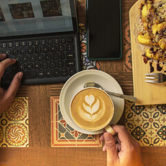 A person savors a latte art coffee and a sweet dessert while typing on a tablet in a charming cafe setting, enjoying a productive day
