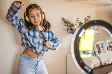 Happy young girl wearing headphones and holding a microphone is singing and dancing, recording a video blog with her smartphone and a ring light