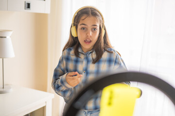 Young girl wearing headphones using a smartphone and a ring light on a tripod to record a video blog, talking and gesturing while standing in her room