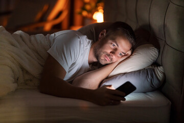 A sleepless man lying in dark bedroom with mobile phone in hands and scrolling endlessly.