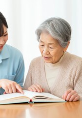 Fototapeta premium A Japanese grandmother sitting at a table, receiving help to read a book from a younger caregiver.