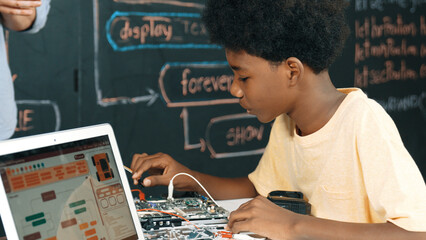 African boy learning to use electronic tool while laptop display engineering code or prompt while sitting in front of blackboard with web develop plan written at STEM technology class. Edification.