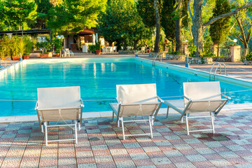 three white desk chairs or chaise lounge with blue swimming pool and hotel green garden on backgeound of vacation summer landscape