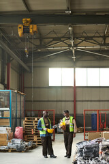 Two male workers of modern factory discussing quality of equipment while standing on aisle between stacks of packed items
