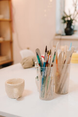 Creative atmosphere for designing ceramic crockery with paintbrushes and handmade ceramic cup on the table. Cozy art studio with sculpting tools.
