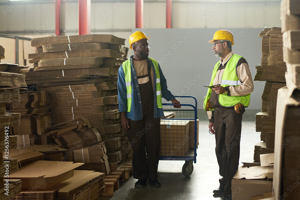 Poster two intercultural engineers having discussion between stacks of cardboard sheets while one of them p
