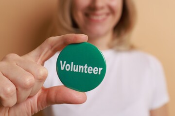 Woman holding button badge with word Volunteer on beige background, closeup
