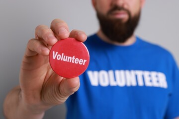 Man holding button badge with word Volunteer on light grey background, closeup