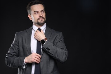 Confident man in classic suit straightening tie on black background. Space for text