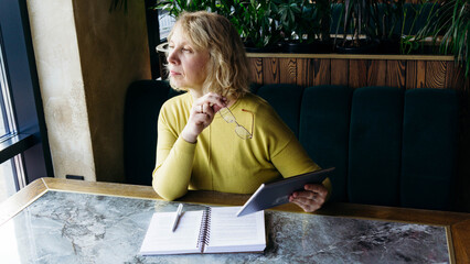 A senior businesswoman works remotely in a restaurant, using a tablet for work while holding...