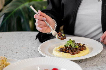 A close-up of a hand holding a fork with savory mushrooms atop creamy mashed potatoes, served on a white plate. The background features a textured gray table and hints of greenery.