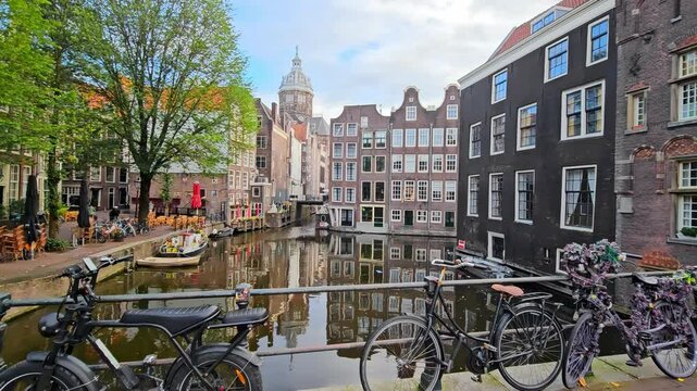Bicycles parked on a bridge overlooking a picturesque canal in Amsterdam, surrounded by historic buildings. A large tree, boats, and outdoor cafes enhance the charm of the scenic view