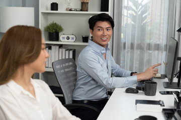 Fototapeta premium Smiling Asian man talking with woman colleague in planning marketing report project on laptop casual day while searching database on working desk at modern home office at side view image. Infobahn.