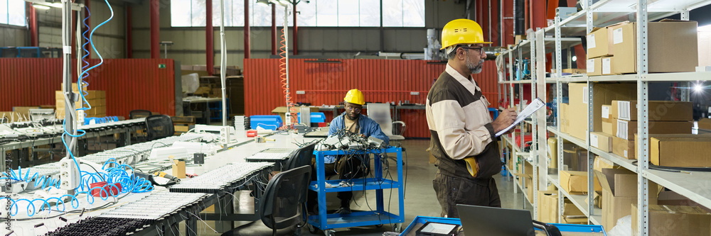 Leinwandbilder header of serious mature worker of factory standing in front of shelves with packed boxes and taking
