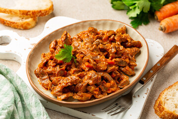 Braised chicken gizzards with onion, carrot, celery, tomato in a rustic terracot plate on a wooden board. Sliced bread.