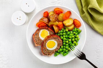 A plate of homemade dinner. Slices of baked meatloaf stuffed with hard-boiled eggs and vegetables, green peas, baby potatoes and carrots.
