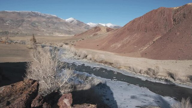 Seething river runs along frozen stone banks. Amazing highland landscape with autumn bare grey forest and snowy mountains against the blue sky. Chaganuzun, Altai