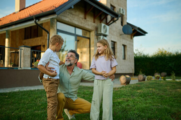 Grandfather crouching in the grass and talking to grandchildren in backyard