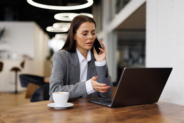 Portrait of young business woman sitting in cafe in front of her laptop and talking on phone