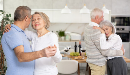Senior men and women doing couple dancing during house party. Two married couples pensioners enjoy move to beat of lounge music in cozy homely atmosphere.
