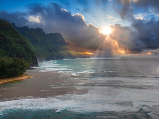 Aerial view of Kauai Island showing Napali Coast cliffs, lush greenery, sandy beach, turquoise waves, and sunlight breaking through clouds.
