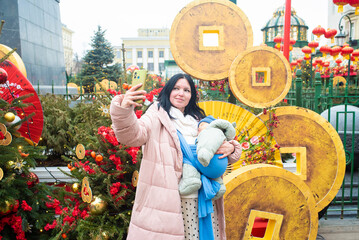 A woman with a sleeping baby in a blue sling takes a selfie on her phone as she stands in front of...