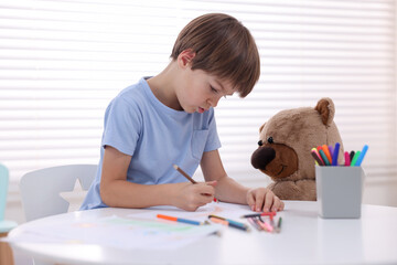 Cute boy drawing near teddy bear at white table in kindergarten