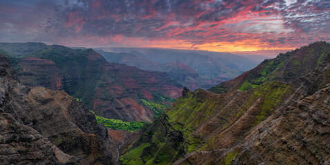 Aerial view of Waimea Canyon's rugged cliffs with green vegetation, red rock formations, and a vivid sunset in pink, orange, and purple hues.