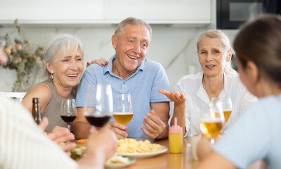 Group of cheerful elderly friends, women and man, gathering around kitchen table, enjoying beer and...