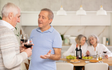 In cozy homely atmosphere, two elderly male friends have withdrawn from their female companions, stand with glass of wine in hands, communicate, discuss tips and tricks, share news