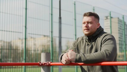 Sportsman takes a break, leaning on a horizontal bar on an outdoor sports field, recovering after...