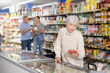 Thoughtful elderly woman buyer going to purchase frozen commodity from cooler in large grocery store