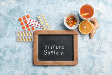 Chalkboard with text IMMUNE SYSTEM, pills, cup of tea, orange and ginger root on color grunge background