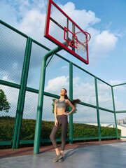 Fototapeta premium Woman posing confidently on a basketball court with a hoop in the background, showcasing athleticism and style against a clear blue sky
