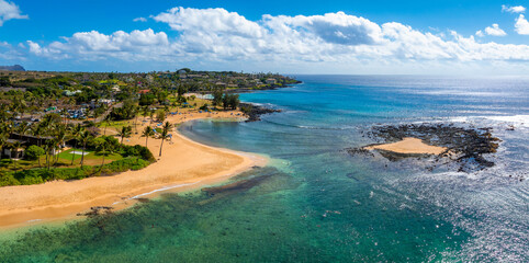 Aerial view of a Kauai beach in Hawaii featuring golden sand, turquoise waters, lush greenery, scattered palm trees, and a rocky outcrop under a blue sky.