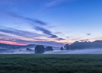 Morning fog is settled over a green Pennsylvania countryside. The sky has shifted from night sky to a dark blue hue with streaks of pink flowing through wispy clouds. © GBS.Images