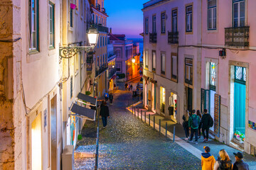Pedestrians walk down a cobblestone hillside street at night with the Tagus River in view a twilight blue hour in the Alfama district of Lisbon Portugal.